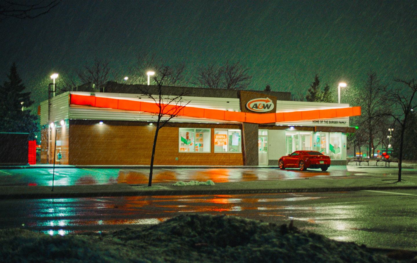 A red car is parked in front of an A&W restaurant at night while it is snowing.