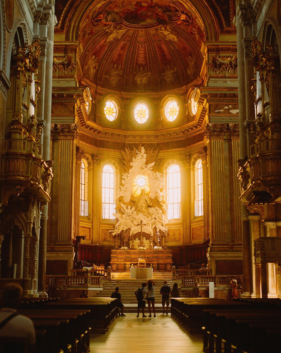 The interior of a grand cathedral features a large altar with a white sculpture and a golden dome.