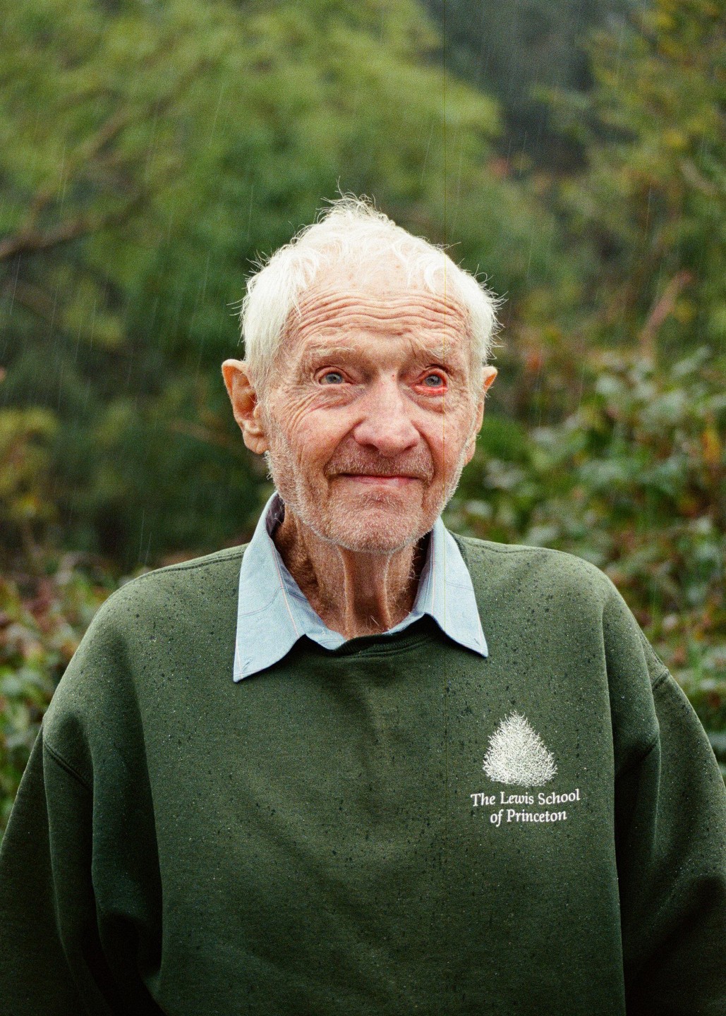 An elderly man with white hair and a green sweatshirt stands in the rain.