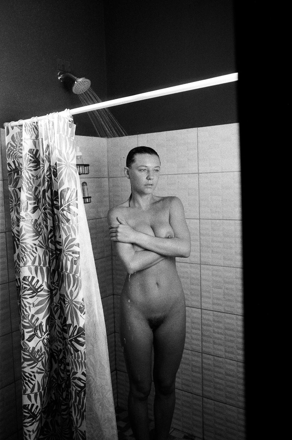 A woman stands naked in a shower with her arms crossed over her chest.