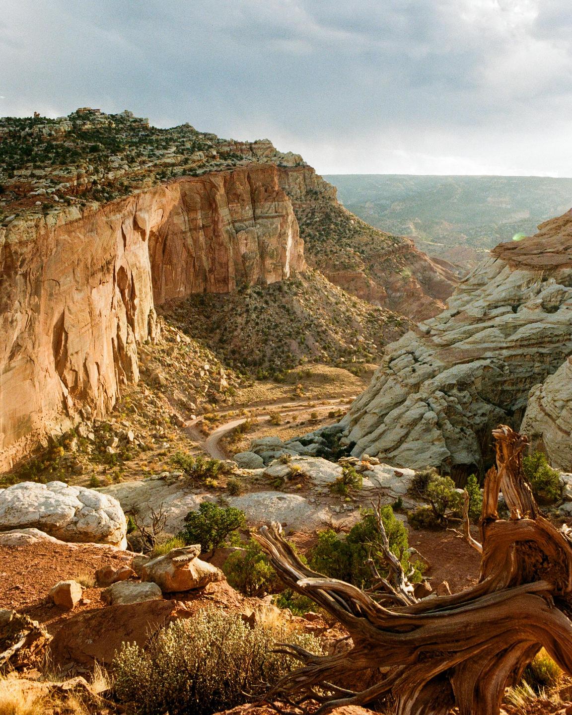 A winding dirt road passes through a canyon with steep rock walls and sparse vegetation under a cloudy sky.