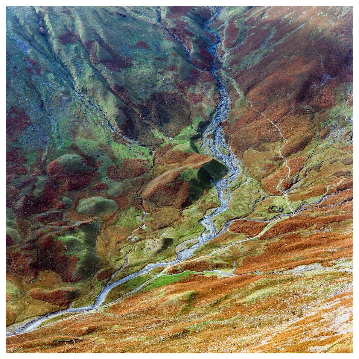 A high-angle landscape shows a winding riverbed through a colorful, rocky, and grassy mountain.