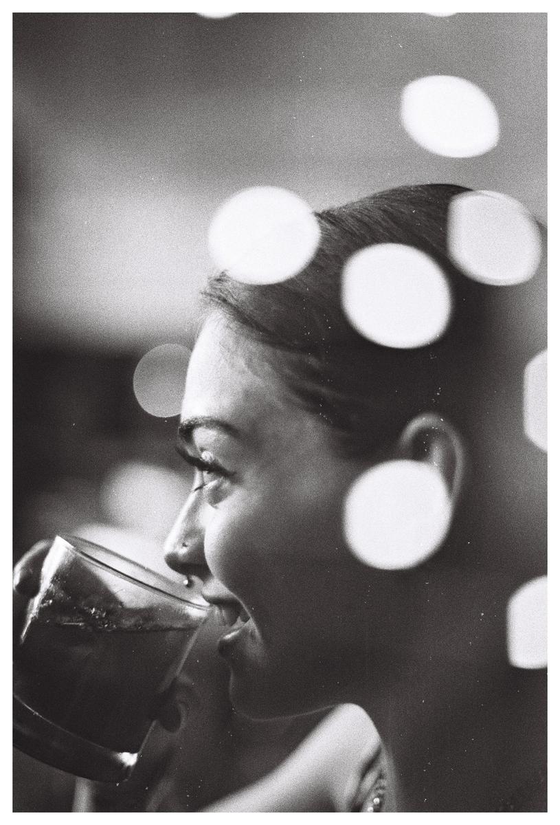A woman with a nose piercing drinks from a glass, with bokeh circles superimposed over her head.
