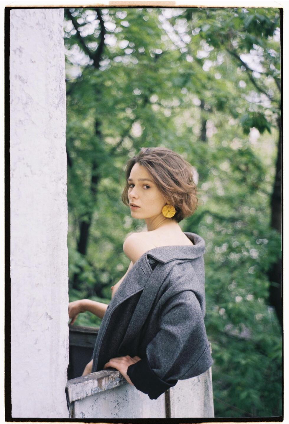 A woman with short hair and a gray coat looks over her shoulder from a balcony with trees in the background.