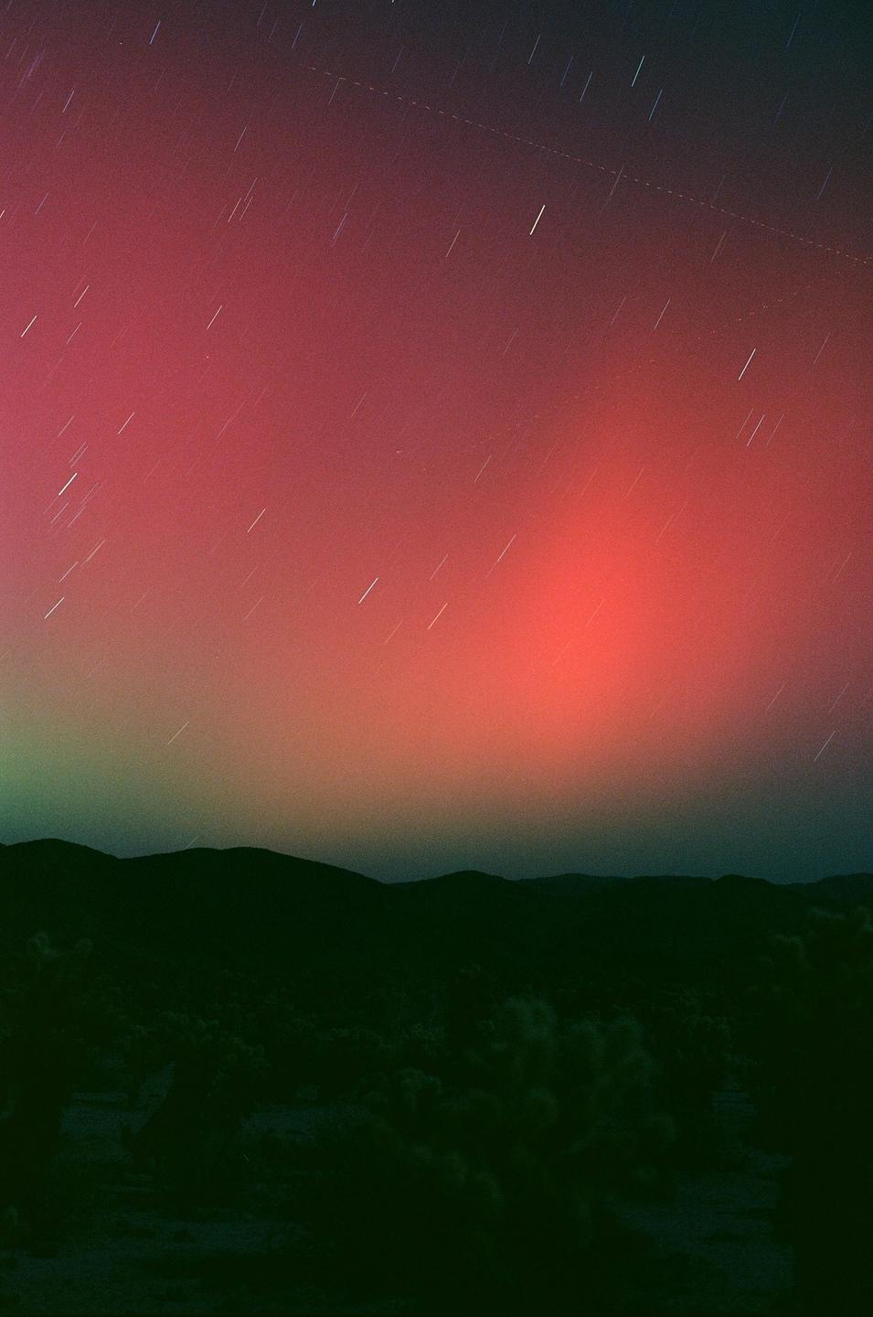 A vibrant red and green aurora glows above a dark mountain range with star trails visible in the sky.