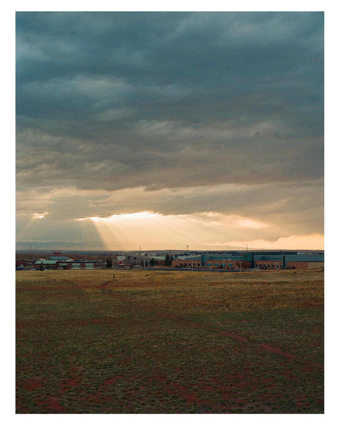 Sunlight streams through dark clouds over a field with buildings in the distance.