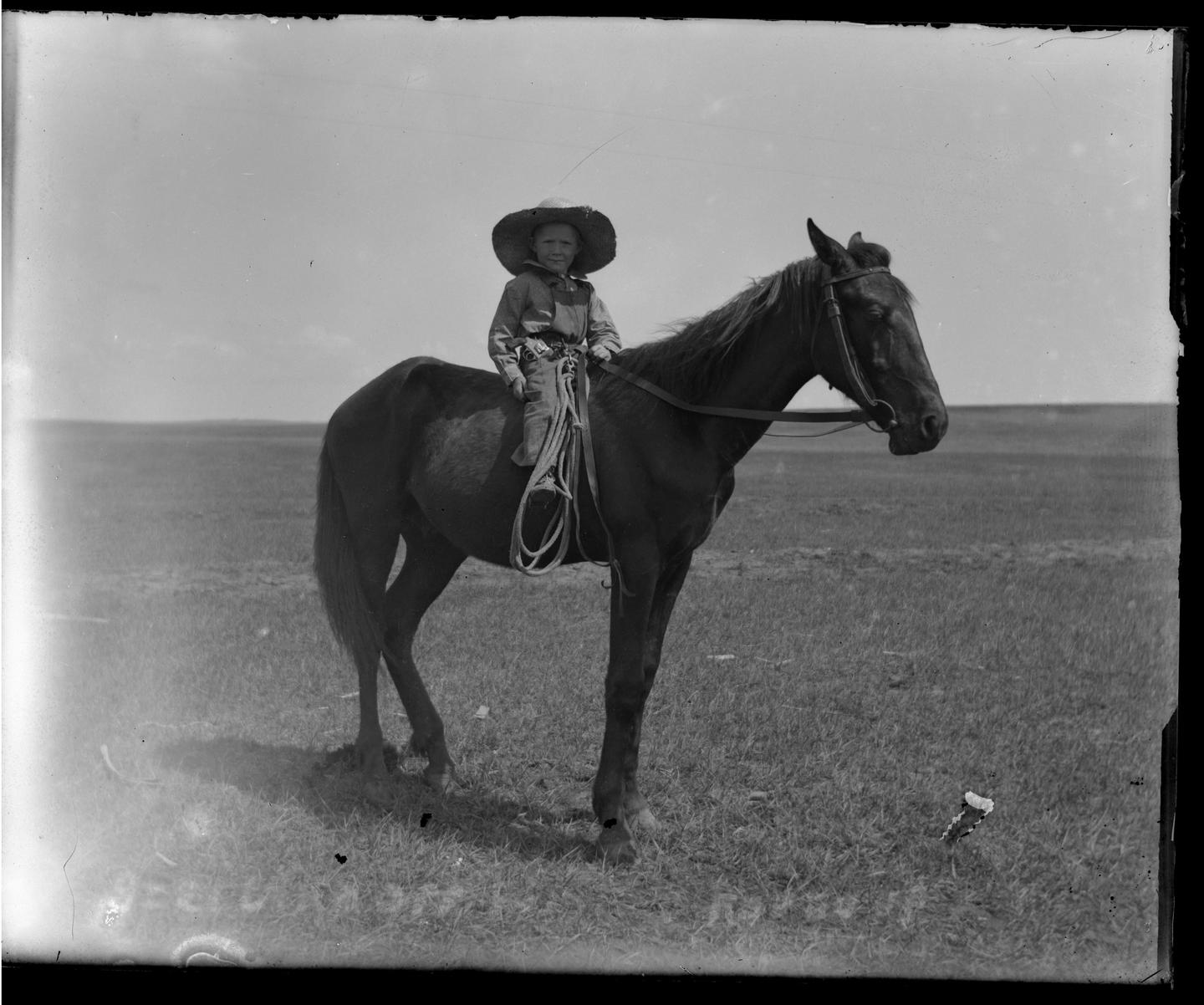 A child wearing a large hat sits atop a horse in a grassy field.