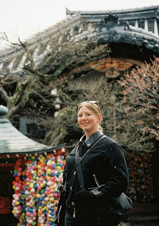 A woman smiles at the camera while standing in front of a traditional building with colorful decorations, as snow falls.
