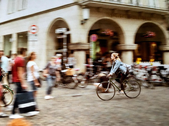 A woman rides a bicycle past a building with arches on a busy street.