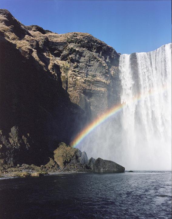 A rainbow arches over a waterfall cascading down a rocky cliff into a body of water.