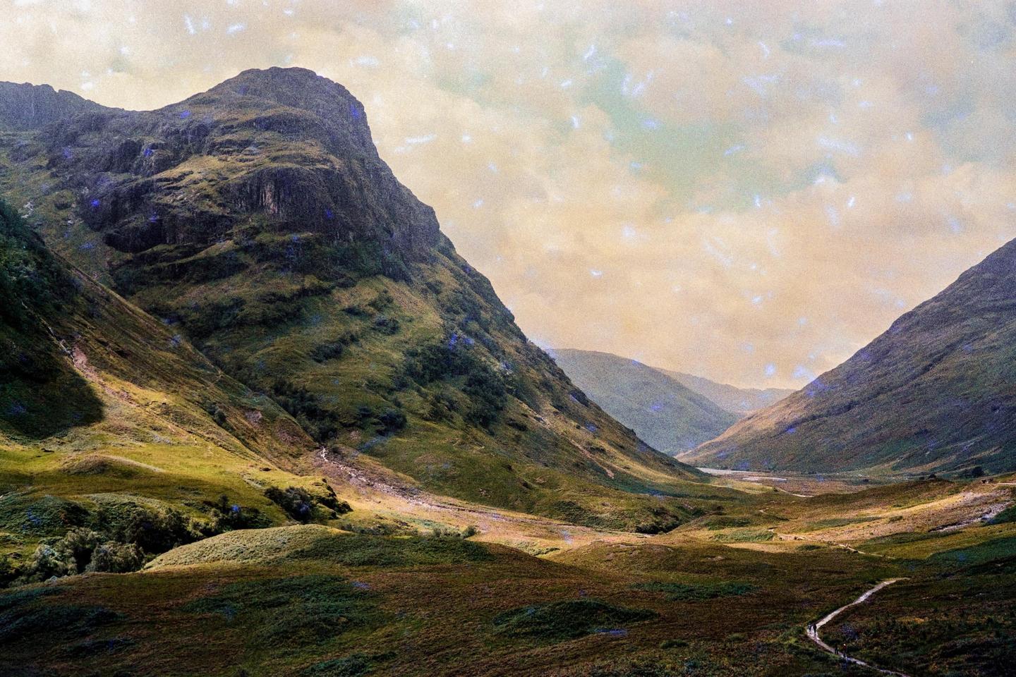 A winding path leads through a vast valley surrounded by mountains under a cloudy sky.