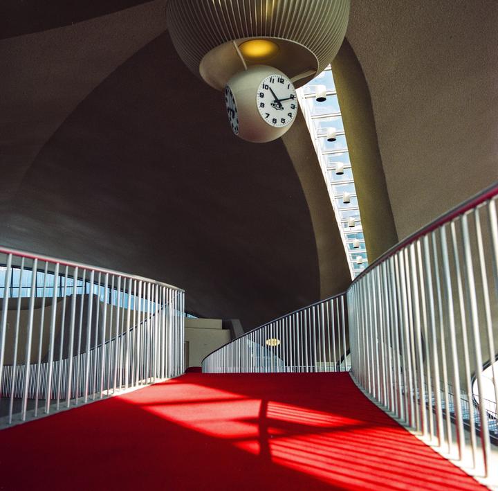 A red carpeted ramp with a metal railing curves under a large clock hanging from a ribbed ceiling.