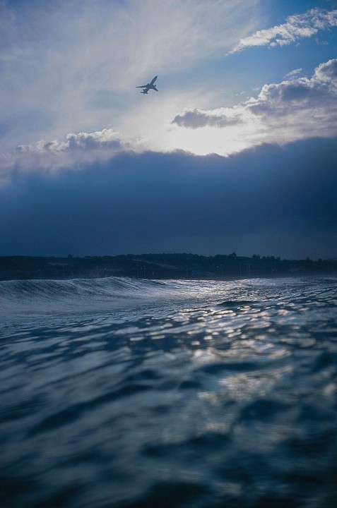 A plane flies over a body of water with a dark, cloudy sky.