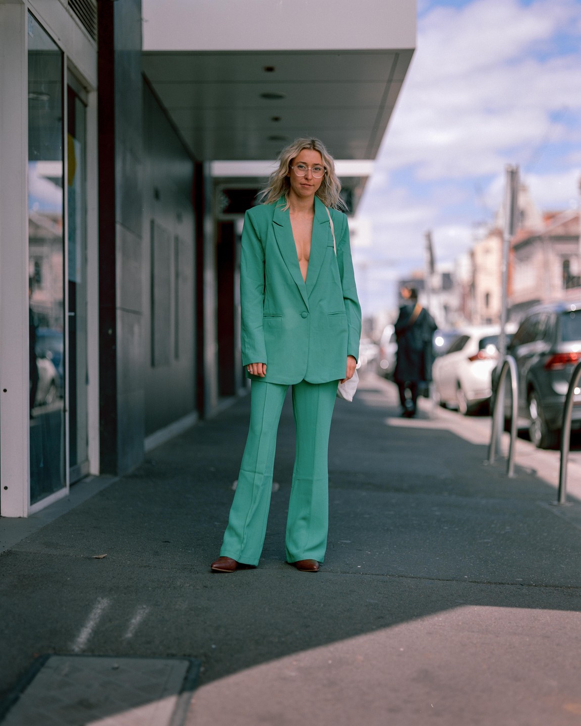 A woman in a green suit stands on a sidewalk next to a building with cars parked in the background.