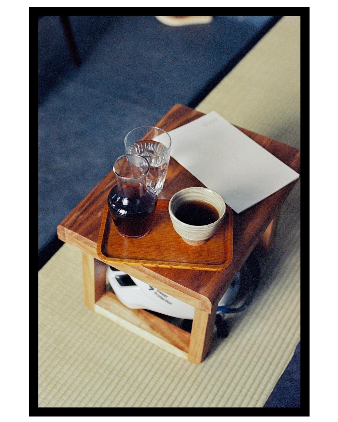 A small wooden table holds a tray with a glass of water, a carafe of coffee, a cup of coffee, and a white paper.