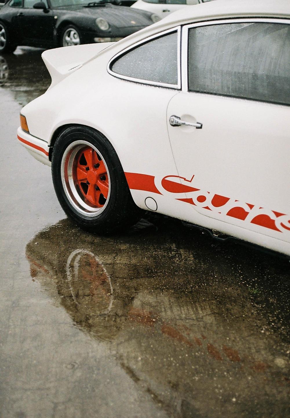 A white Porsche with red accents is parked on a wet surface, reflecting in a puddle.