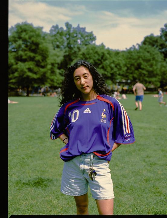 A woman wearing a blue soccer jersey and denim shorts stands in a grassy park.