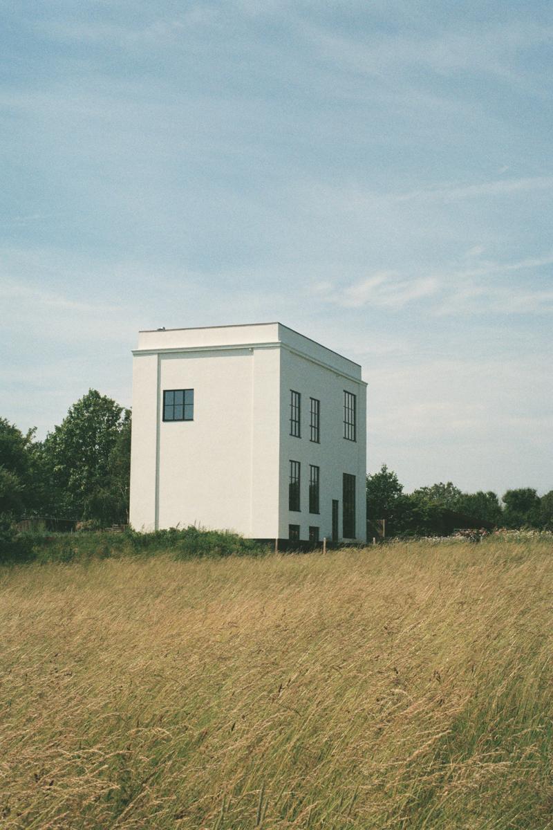 A white rectangular house with dark windows stands in a field of tall dry grass under a blue sky.