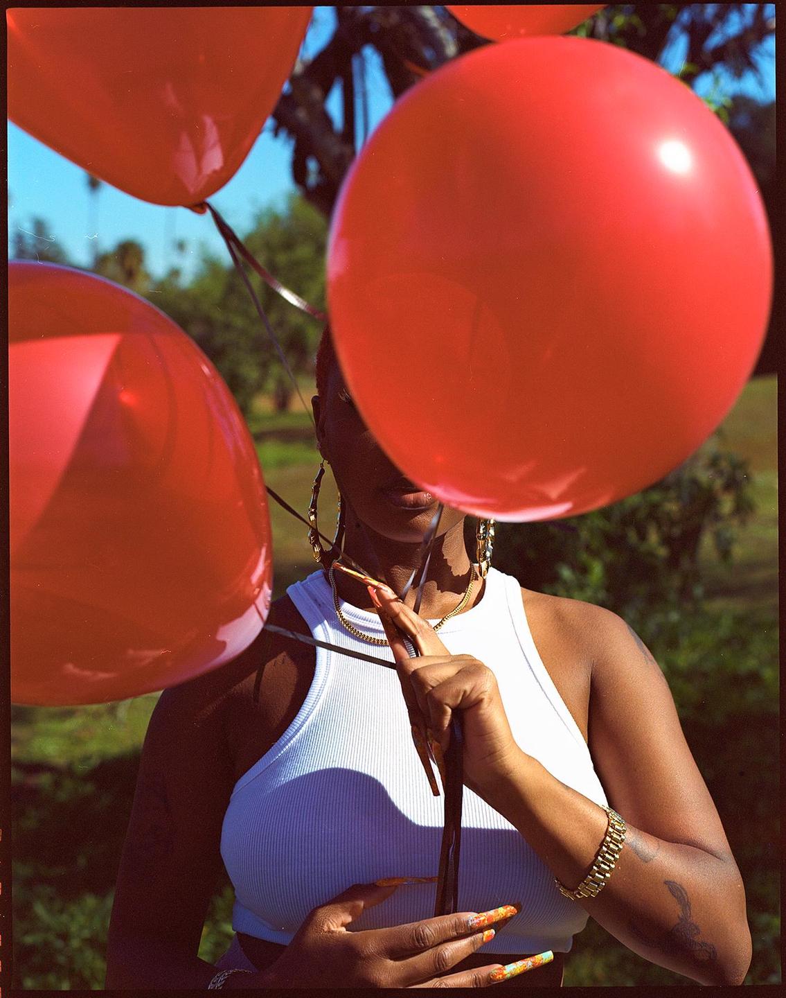 A woman holds red balloons that obscure her face in a sunlit outdoor setting.
