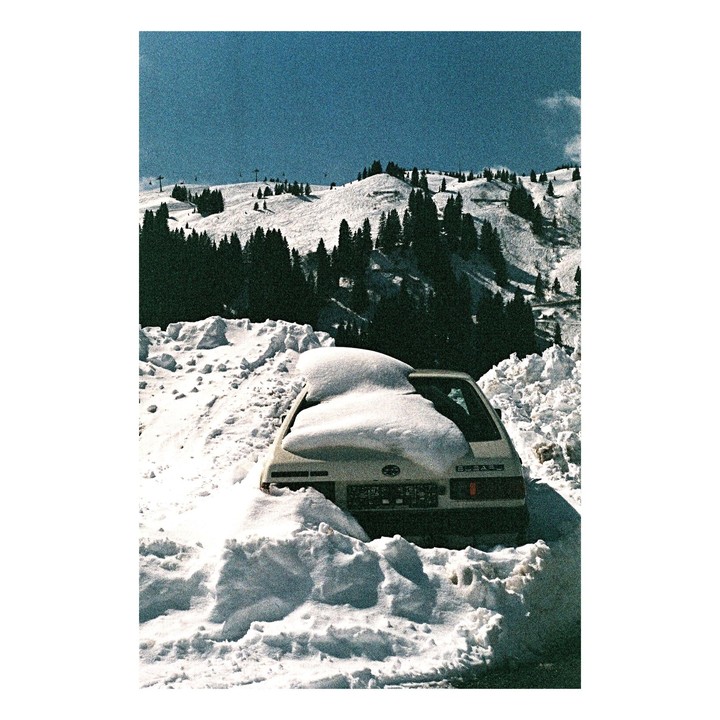 A car is buried in snow with a mountain covered in pine trees and snow in the background.
