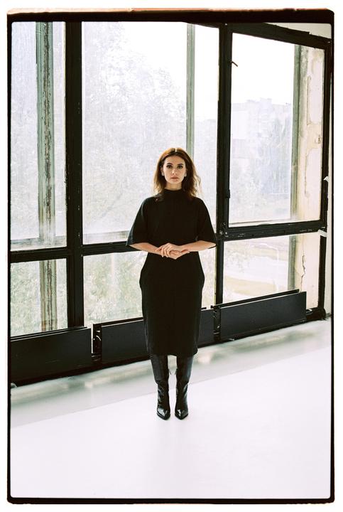 A woman in a black dress and boots stands in front of a large window.
