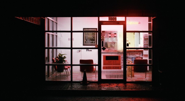 A storefront is illuminated with a red glow at night, revealing a waiting area with chairs and a plant inside.