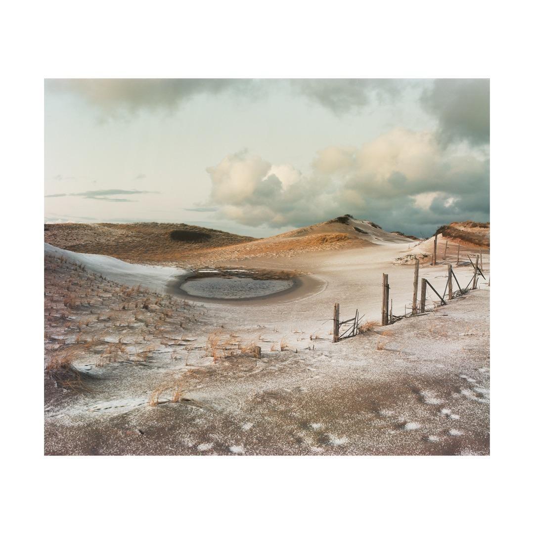 A frozen pond sits in a snowy, hilly landscape with a broken fence under a cloudy sky.