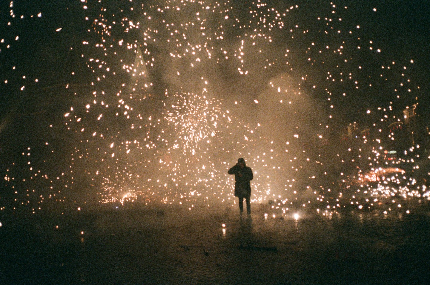 A person stands in the middle of a street at night, surrounded by fireworks and smoke.