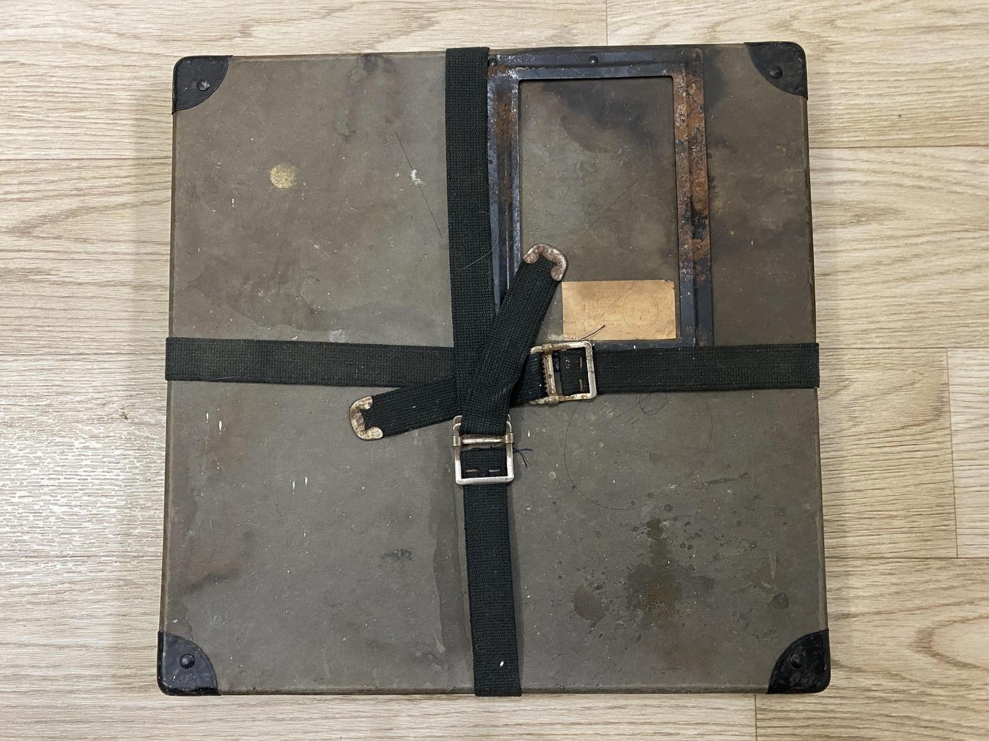 A dark, rectangular box with metal corners and a black strap is resting on a wooden floor.