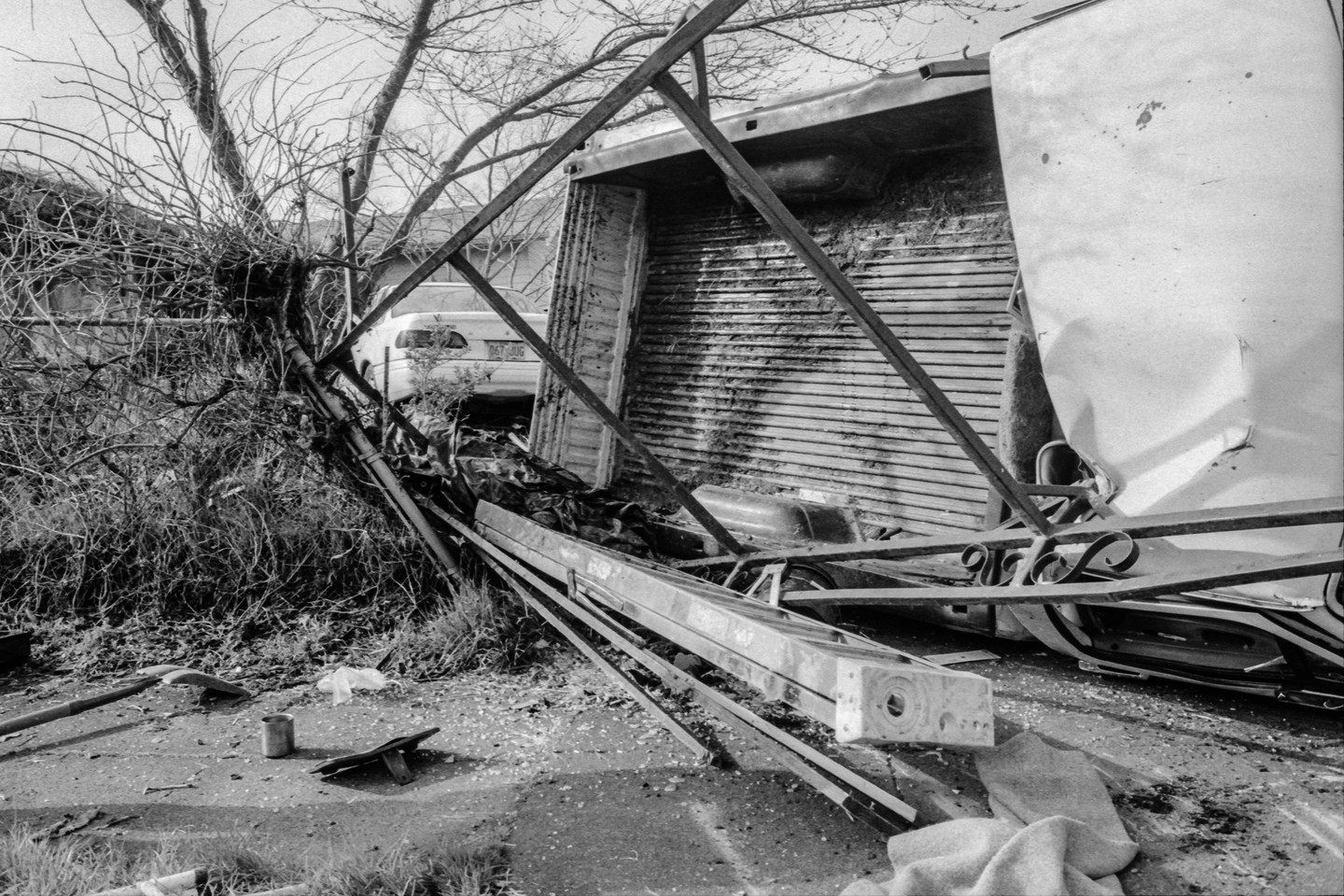 A black and white image shows a scene of destruction with a fallen tree, a damaged truck bed, and debris scattered on the ground.