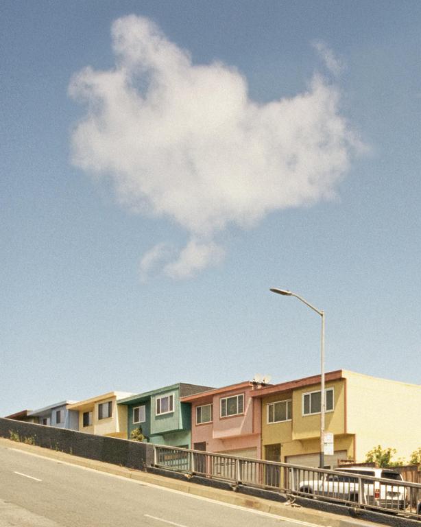 A large cloud floats above a row of colorful houses on a hill next to a street.