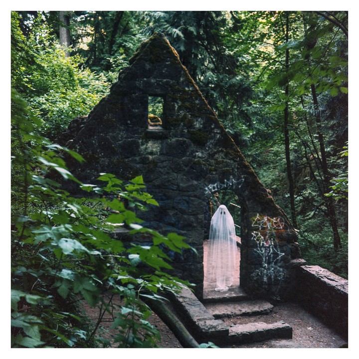 A person dressed as a ghost stands in the doorway of a stone structure in a forest.