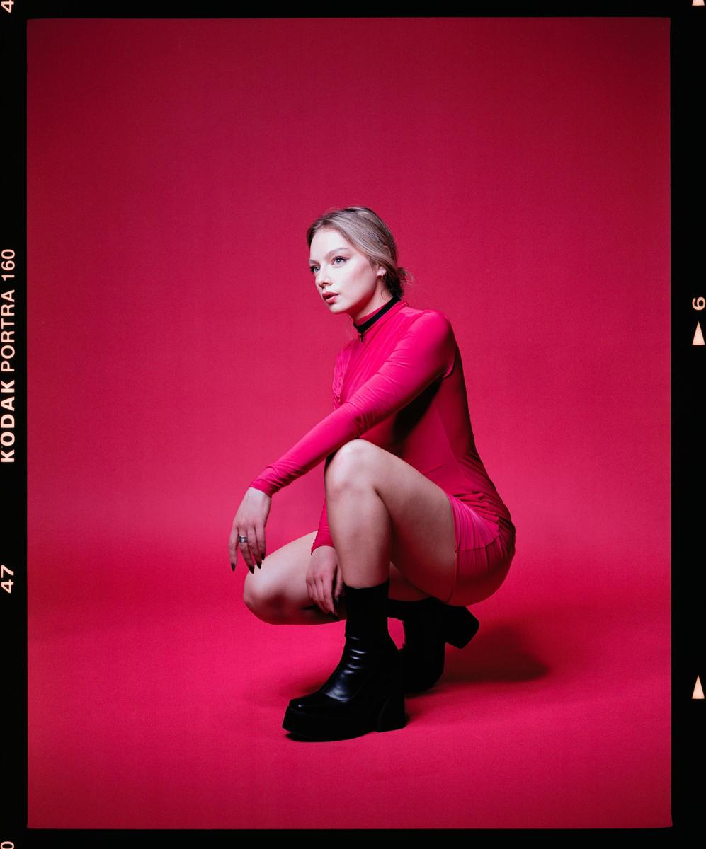 A woman in a pink dress and black boots crouches against a solid pink background.