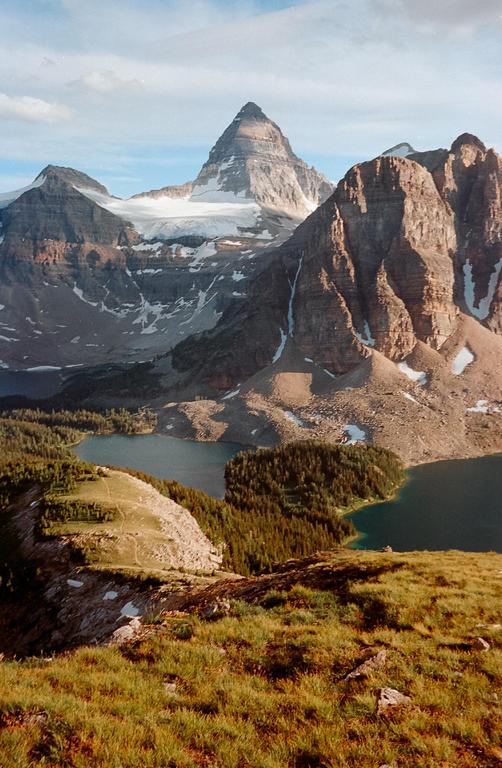 A mountain range with snow-capped peaks overlooks a valley with two lakes and a dense forest.