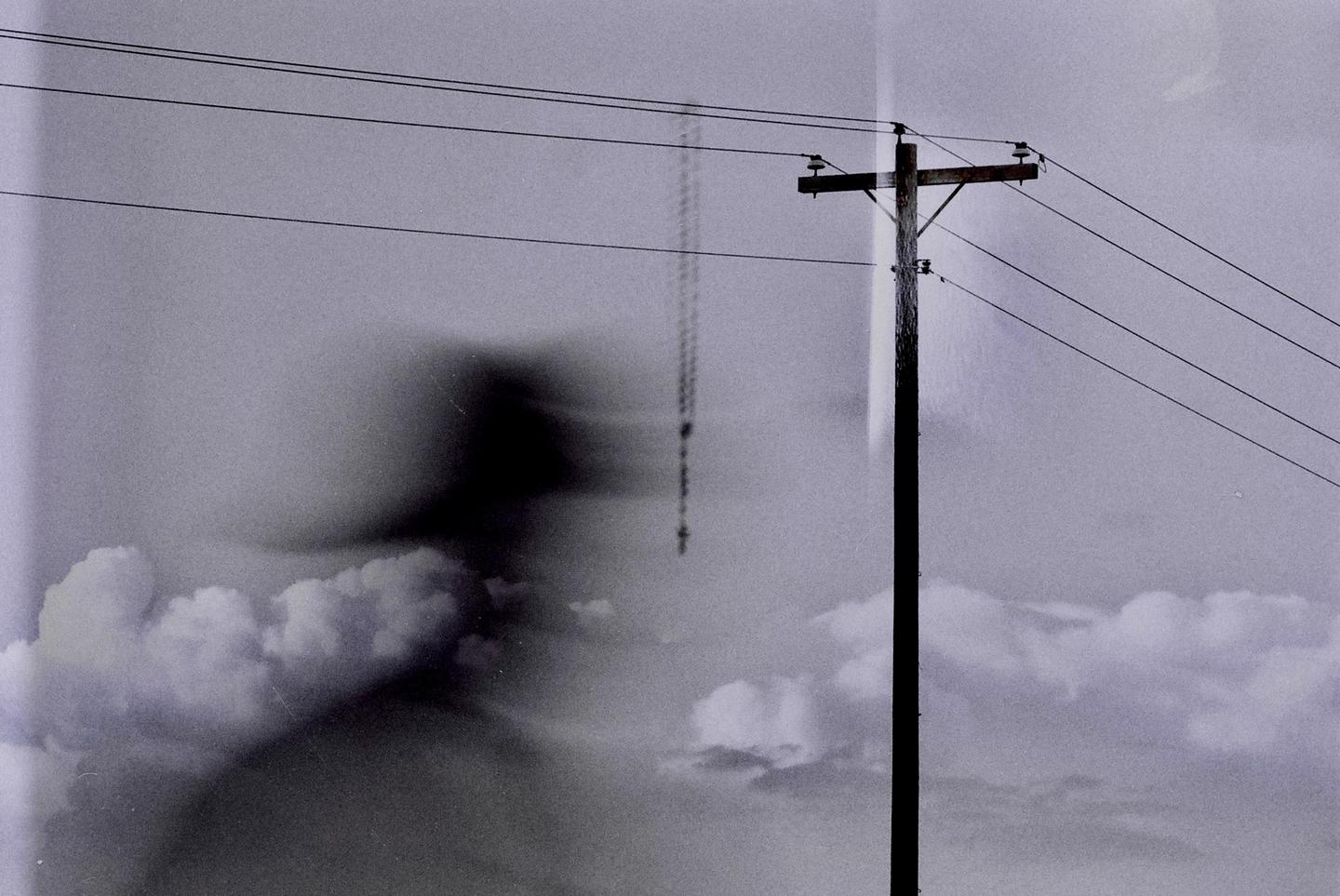 A telephone pole with wires stands against a cloudy sky with a faint, dark, ghost-like figure on the left.