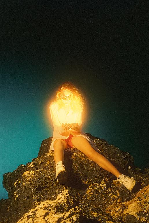 A woman with glowing hair and sunglasses sits on a rocky outcrop against a dark sky.