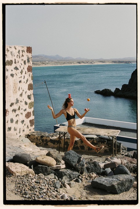 A woman in a bikini balances a can on her head and fruit in her hands while standing on a rocky cliff overlooking the ocean.