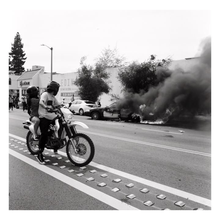 A man and woman on a motorcycle drive past a burning car on a street.
