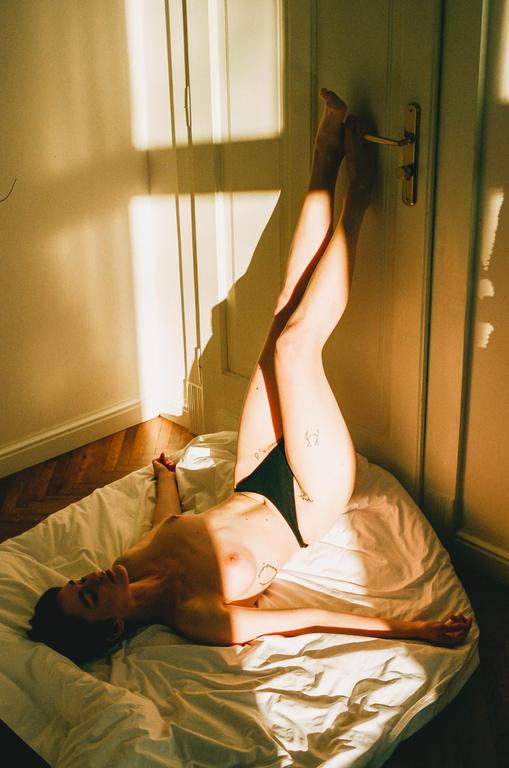 A woman lies on a white sheet with her legs up against a door, illuminated by golden hour sunlight.