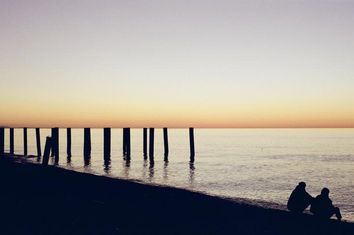 Two people sit on a beach at dusk with a row of pier pilings extending into the water.