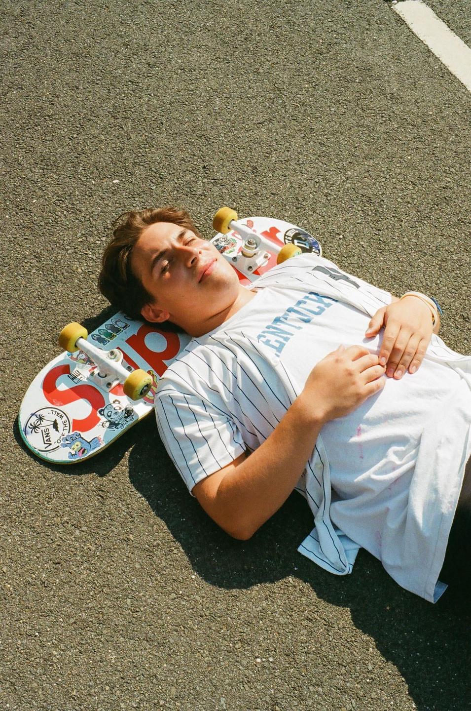 A young man with closed eyes is lying on a skateboard on an asphalt surface.