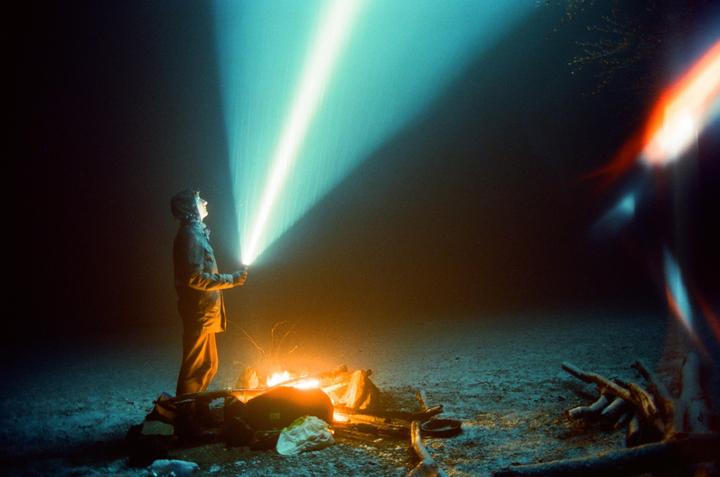 A person in a hooded jacket stands by a campfire at night, shining a bright light into the dark sky.