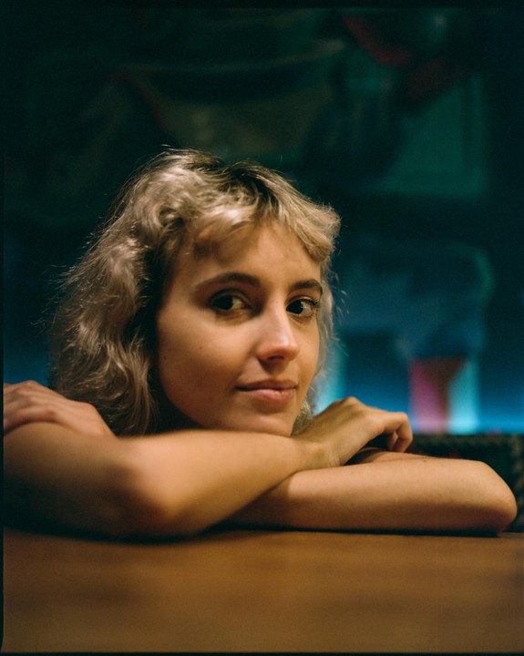 A woman with blonde hair rests her arms on a wooden table, looking towards the viewer.