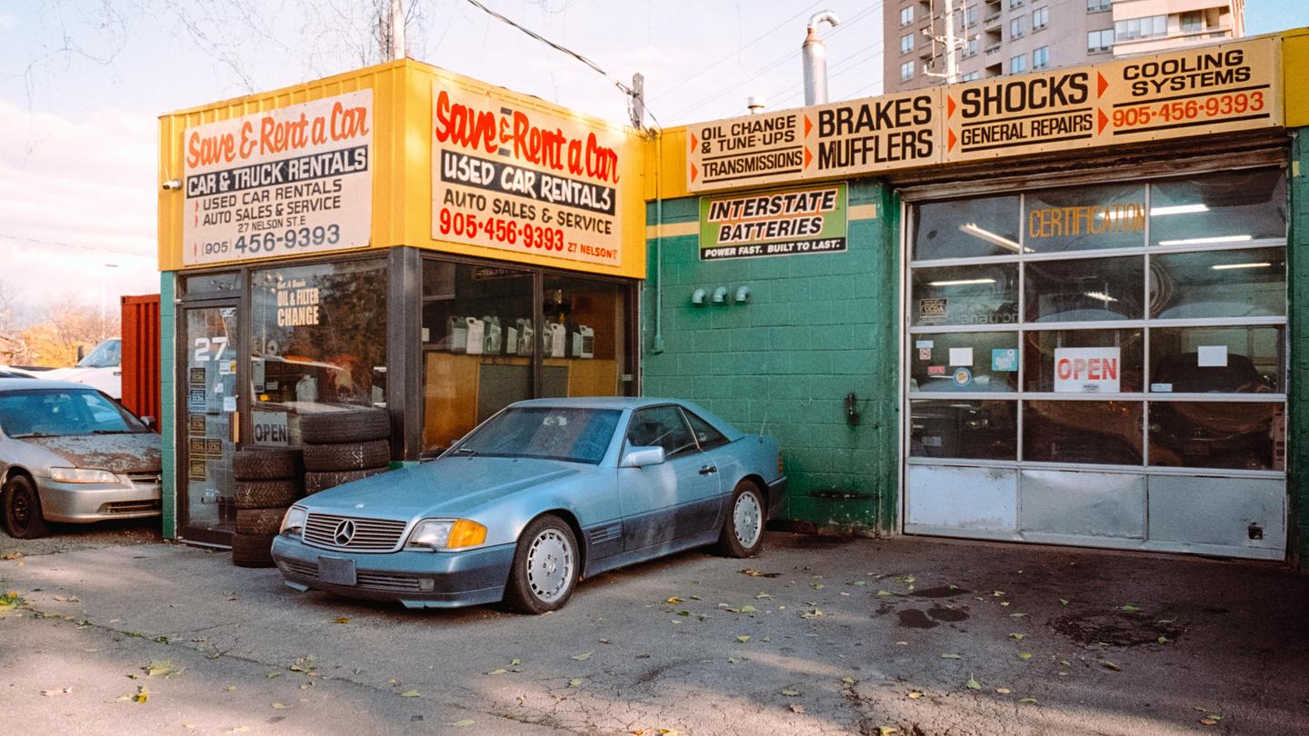 A blue car is parked in front of a car rental and repair shop with yellow and green signage.