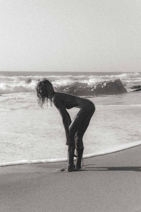 A woman bends over on a sandy beach with waves and rocks in the background.