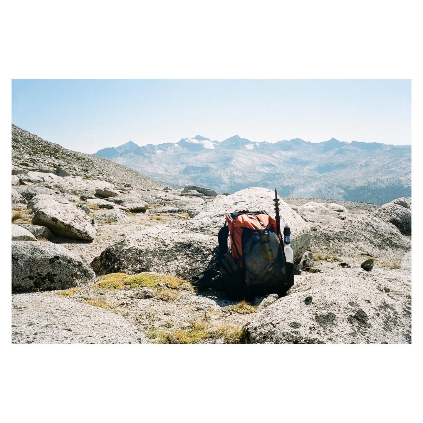 A backpack sits on a rocky mountain with snow-capped peaks in the distance.