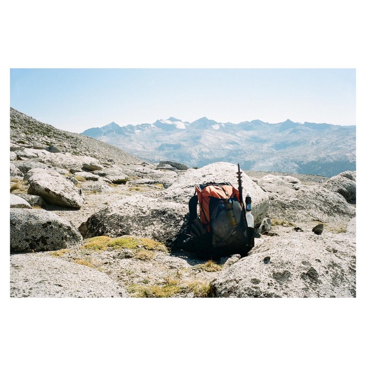 A backpack sits on a rocky mountain with snow-capped peaks in the distance.