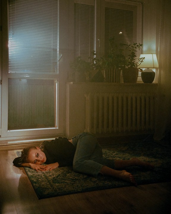 A woman lies on a patterned rug on the floor next to a window with blinds, with plants and a lamp on the windowsill.
