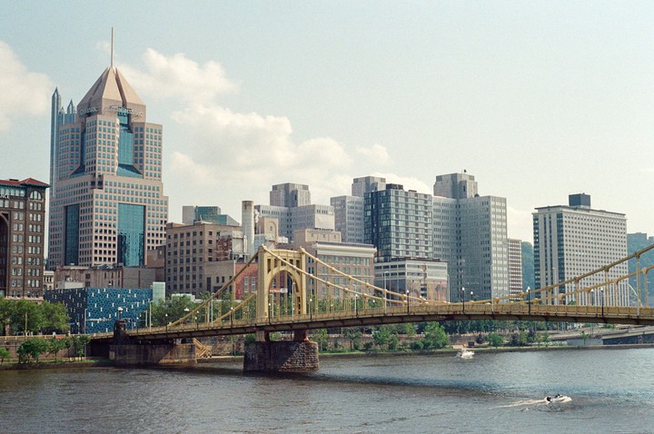A cityscape with a prominent skyscraper and a bridge spanning a river, with boats on the water.
