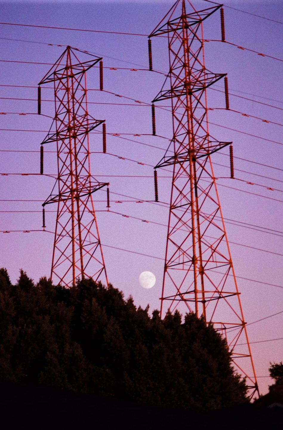 Two power line towers stand against a purple sky with a full moon visible above a line of dark trees.